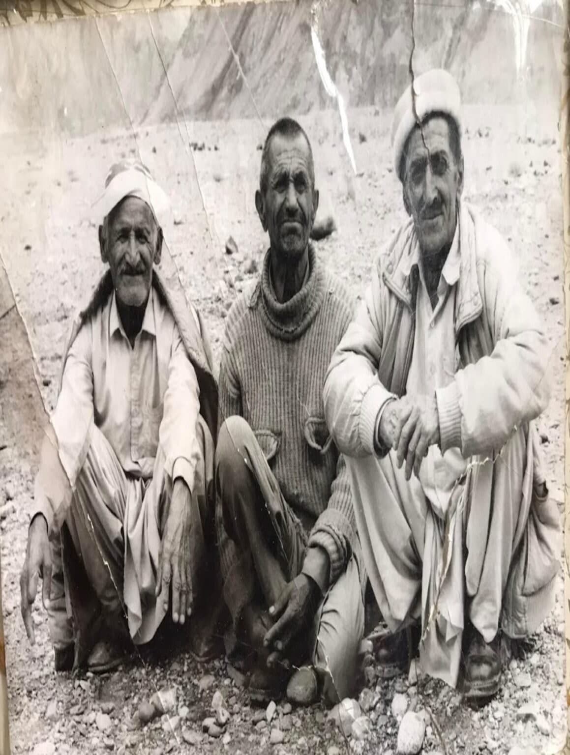 An archival black-and-white photograph of three elders seated on rocky ground