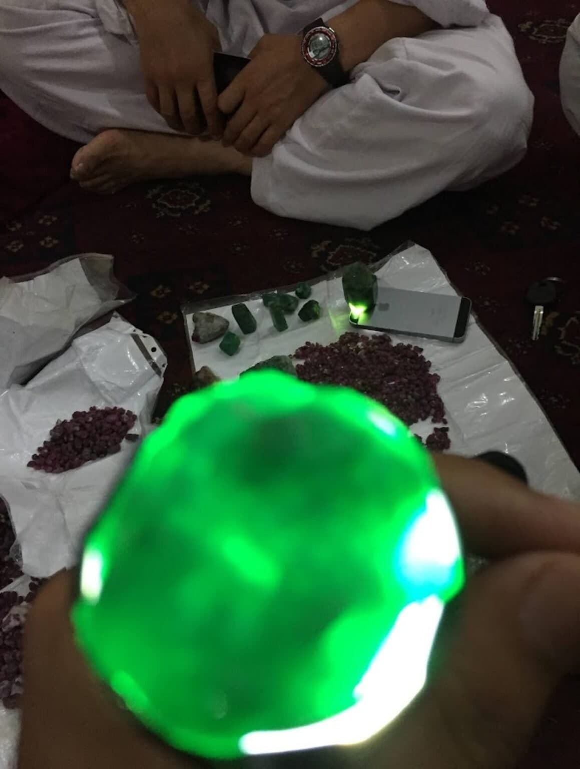 A green crystal held up backlit during a specimen sourcing session, with ruby crystals nearby