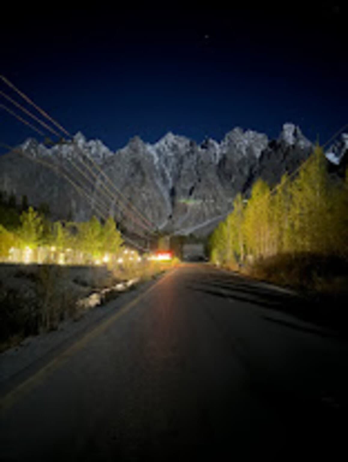A mountain road at night lit by streetlights, with peaks visible under starlight