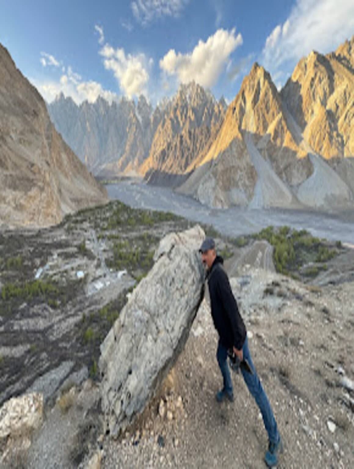 A man leaning on a large rock overlooking the Passu Cones and river valley below